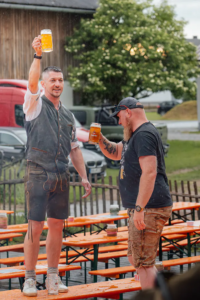 Zwei Männer mit Maßkrügen stoßen im Regen zwischen nassen Festbänken auf Zoiglbier an.