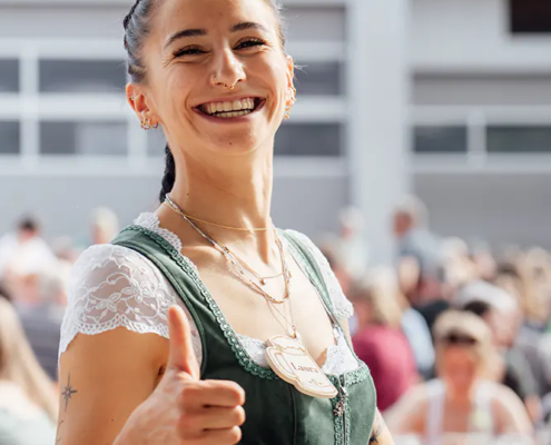 Laura Bedienung im grünen Dirndl zeigt Daumen hoch und lächelt zwischen den Festbänken.