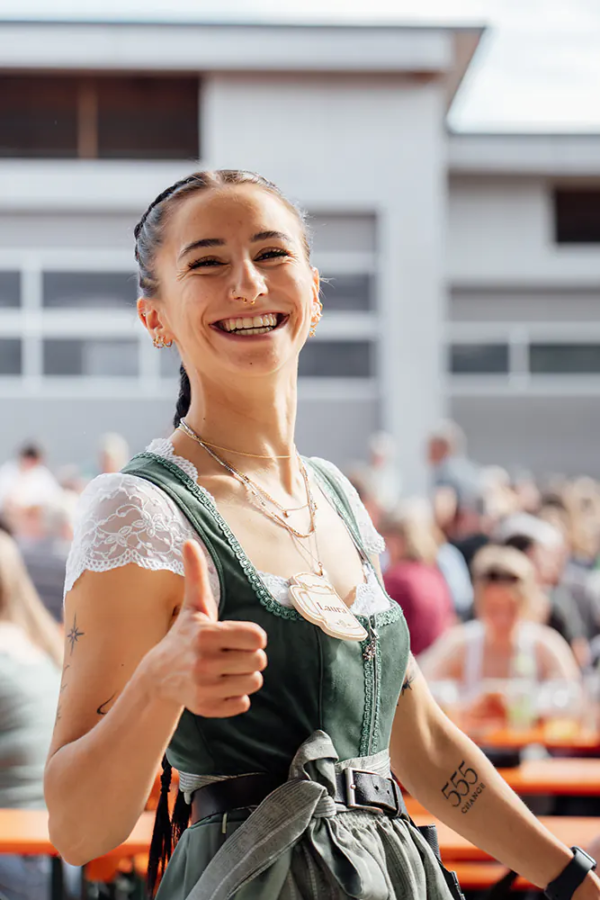 Laura Bedienung im grünen Dirndl zeigt Daumen hoch und lächelt zwischen den Festbänken.