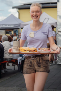 Bedienung im Festshirt und Lederhosen serviert Brotzeit und Salat auf einem Tablett.